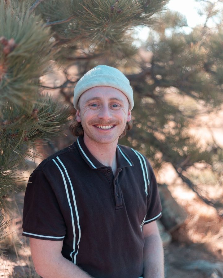 Sam smiling in a light mint beanie and a dark polo with pale stripes, in front of a pine tree
